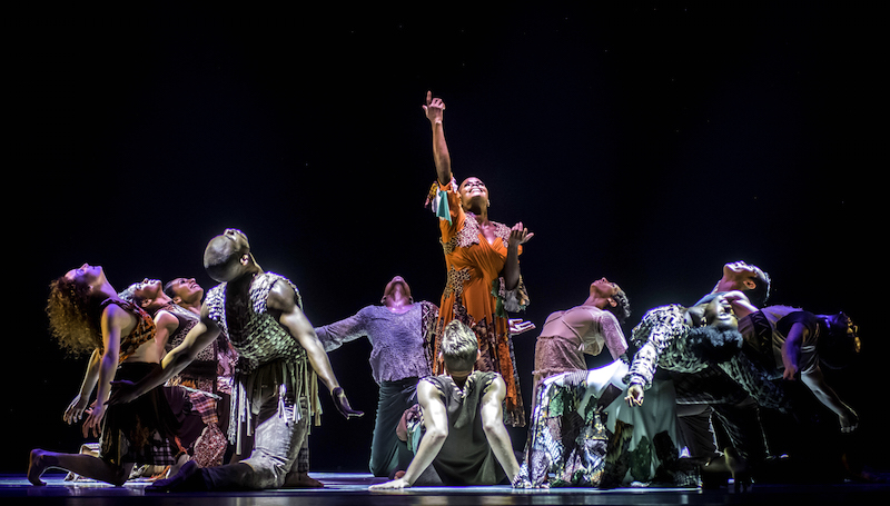 A female dancer in a bright red dress lifts her arms celestially to the sky while a large group of dancers kneel beside her with her heads back